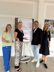 Drei Frauen und ein Mann lächeln oder lachen in die Kamera, manche gestikulieren. Hinter ihnen eine Flipchart mit Handschrift: "Wirkung ist veränderbar … JETZT"