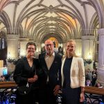 Zwei Frauen und ein Mann stehen auf einer Art Balkon, im Hintergrund und dahinter eine imposante Säulenhalle im Rathaus Hamburg. Unten ist die Fläche fast komplett mit Menschen gefüllt.