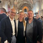 Eine Frau steht zwischen zwei Männern inmitten des Publikums aus dem ersten Foto. Man erkennt eine imposante Säulenhalle, im Rathaus Hamburg.