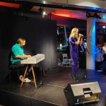Eine Frau steht auf einer kleineren Bühne am Mikrofon und singt. Hinter ihr ein Mann an einem kleinen weißen Piano.