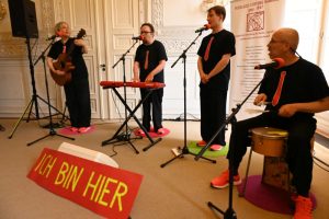 Eine Band mit einer Frau und drei Männern live. Instrumente: Gitarre, Keyboard, Trommel. Alle tragen neonorangene Sneaker und dunkle T-Shirts mit aufgedruckten roten Krawatten