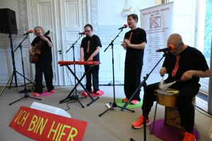 Eine Band mit einer Frau und drei Männern live. Instrumente: Gitarre, Keyboard, Trommel. Alle tragen neonorangene Sneaker und dunkle T-Shirts mit aufgedruckten roten Krawatten