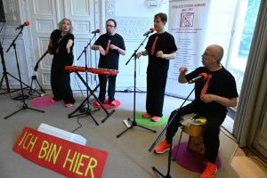 Eine Band mit einer Frau und drei Männern live. Instrumente: Gitarre, Keyboard, Trommel. Alle tragen neonorangene Sneaker und dunkle T-Shirts mit aufgedruckten roten Krawatten