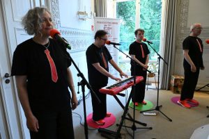 Eine Band mit einer Frau und drei Männern live. Instrumente: Gitarre, Keyboard, Trommel. Alle tragen neonorangene Sneaker und dunkle T-Shirts mit aufgedruckten roten Krawatten