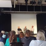 Publikum von hinten, vorne auf der Bühne 3 Personen im Gespräch.