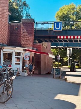 Eine U-Bahn-Haltestelle aus rotem Backstein, man erkennt die U-Bahn-Brücke. Darunter eine Eisdiele. Die Sonne scheint.