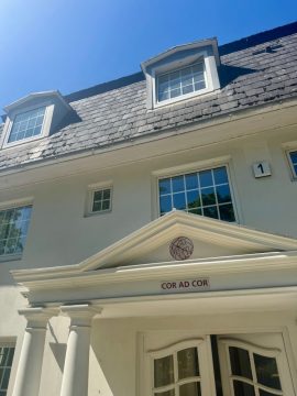 Ein Teil der Fassade eines villenartigen weißen Hauses. Die Sonne scheint schön. Die Doppel-Türe ist altmodisch aber schick. Über der großen Türe des Hauses ein Schild mit der Aufschrift "Cor ad cor".